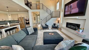 A spacious living area with a large gray sofa, a fireplace, a TV mounted on the wall, and a kitchen in the background.