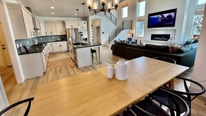 Una cocina moderna con una isla central, un comedor con mesa y sillas de madera, una sala de estar con sofá y televisor, y una escalera que sube a un nivel superior.