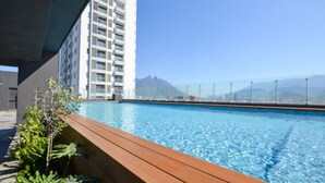 Une piscine dotée d’une terrasse en bois, entourée d’un grand immeuble et offrant une vue sur la montagne.