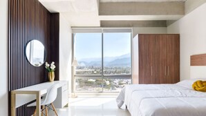 Une chambre moderne avec un grand lit, un bureau, un miroir et une vue sur le paysage urbain, visible à travers une grande fenêtre.
