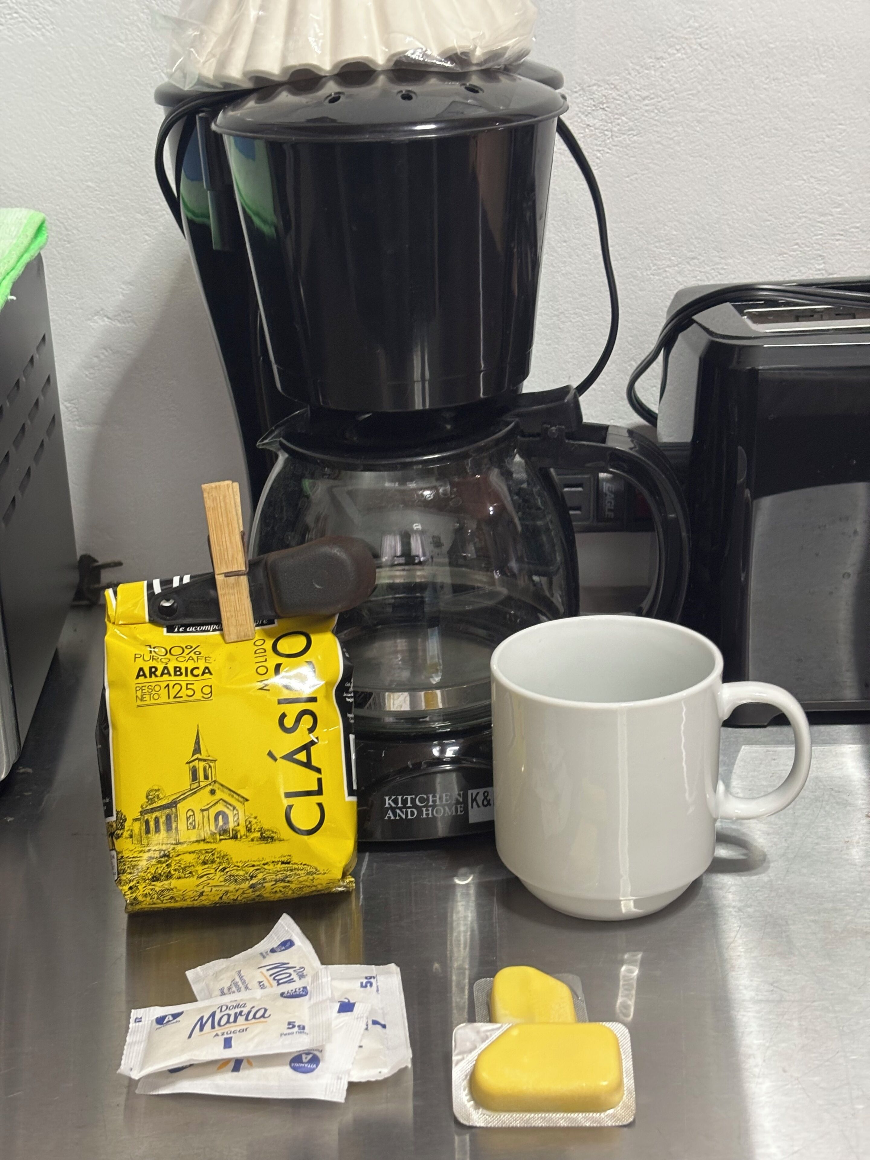 Un comptoir de cuisine avec une machine à café, un paquet de café, une tasse blanche, des sachets de sucre et un sachet de beurre.