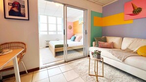 Ein modernes Wohnzimmer mit einer Couch, einem kleinen Tisch, einem Korbstuhl und einer Schiebeglastür, die zum Schlafzimmer führt.