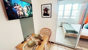 Ein Zimmer mit Esstisch für zwei Personen, einem an der Wand befestigten Fernseher, einem gerahmten Bild und einer Glasschiebetür, die zu einem Bett führt.