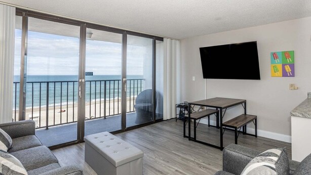Un salon moderne avec un canapé, un pouf et une table, un téléviseur à écran plat fixé au mur, et un balcon offrant une vue sur la plage.