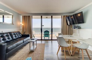 Ein modernes Wohnzimmer mit einer Ledersofa, einem Esstisch mit Stühlen, einem Flachbildfernseher und einem Balkon mit Blick auf den Strand.