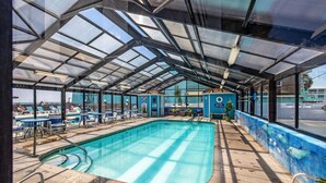 Piscina techada con sillas y mesas de descanso, un mural azul y una vista despejada del cielo.