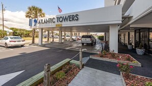 Entrada de hotel con un letrero que dice 'ATLANTIC TOWERS', zona de estacionamiento techada y un acceso ajardinado con macizos de flores.