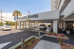 Entrada de hotel con un letrero que dice 'ATLANTIC TOWERS', zona de estacionamiento techada y un acceso ajardinado con macizos de flores.