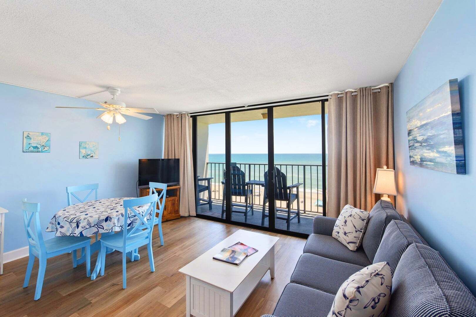 A room with a balcony, a dining table with chairs, a sofa, a TV, and artwork on the walls.