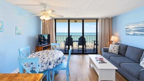 Una habitación con balcón, televisor, sofá, mesa y sillas.