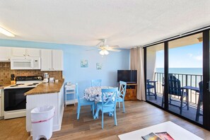 A kitchen with a microwave, stove, and sink, a dining area with a table and chairs, and a balcony with a view of the ocean.