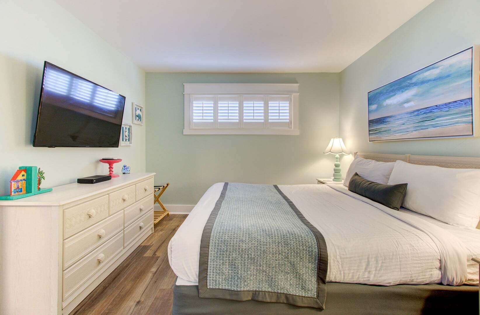 A bedroom with a bed, a television, a window, and a painting on the wall.