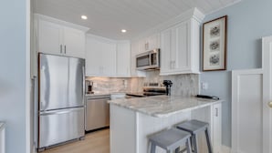 A modern kitchen with white cabinetry, stainless steel appliances, a marble countertop, and a small island with bar stools. - Oceanfront retreat in Carolina Beach 0910 (Carolina Beach)