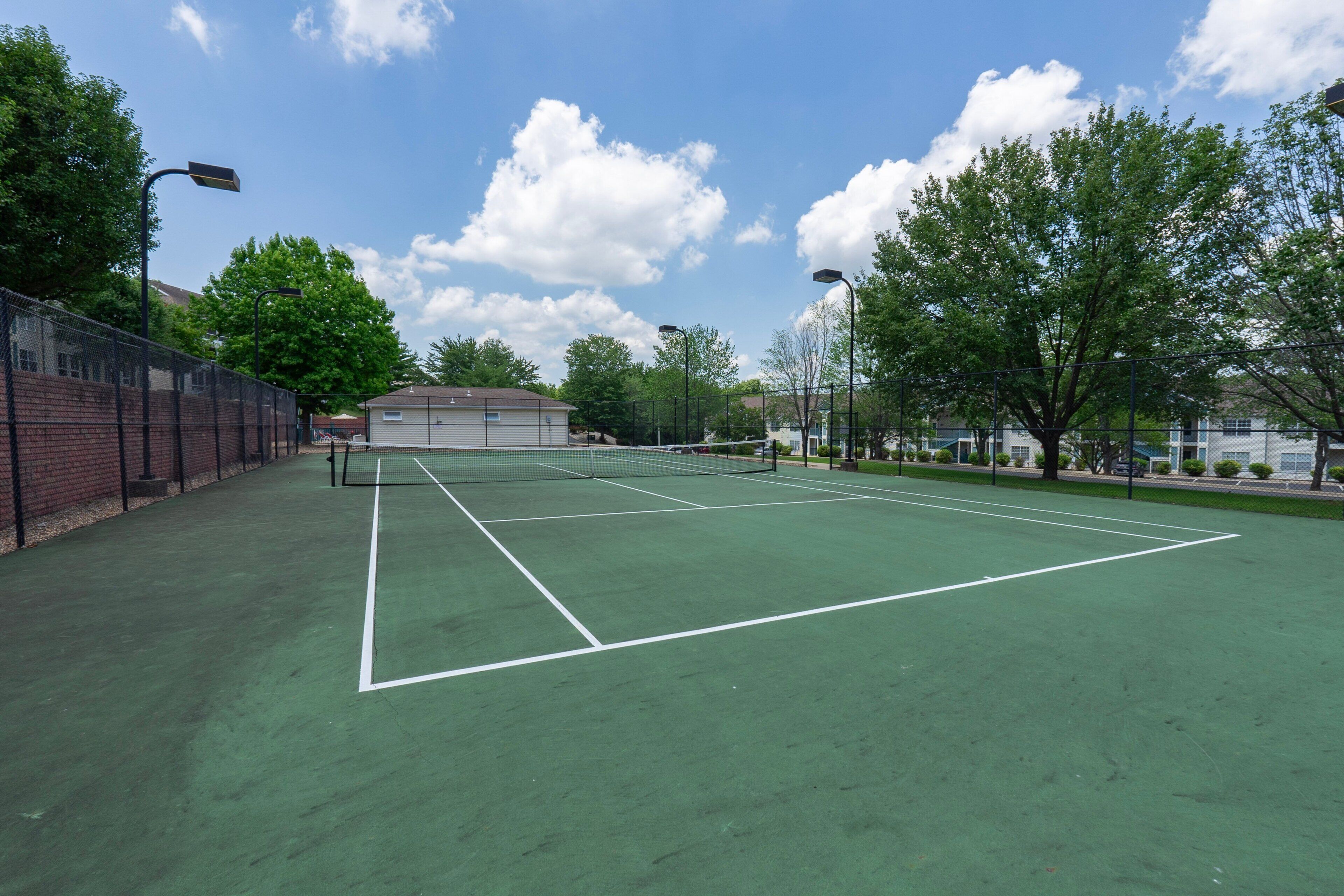 A tennis court with a net, surrounded by a fence, trees, and buildings in the background.