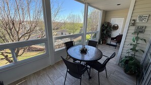 Eine überdachte Terrasse mit Tisch und Stühlen, großen Fenstern, einem Deckenventilator und Blick auf Bäume und ein Haus.