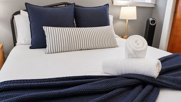 A neatly made bed with a striped pillow, a rolled white towel, and a dark blue blanket. - Elegant Harvard Free Parking • Prime Location, 2 Bed 1 Bath (Cambridge)