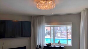 Ein Wohnzimmer mit Fernseher, einer Kristallleuchter, Blick auf einen Pool und einem Klavier.