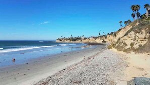 Ein Strand mit Kieselsteinen, einem klaren Himmel und Palmen auf einem Hügelhang.