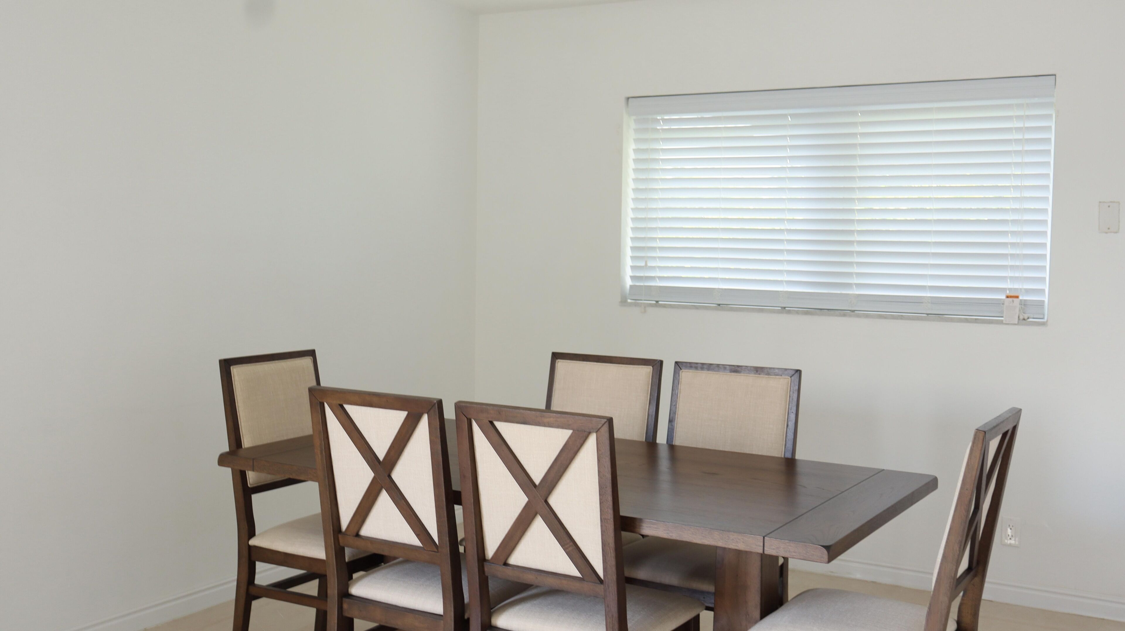 A dining area with a rectangular table and four chairs, a window with blinds, and a plain white wall.