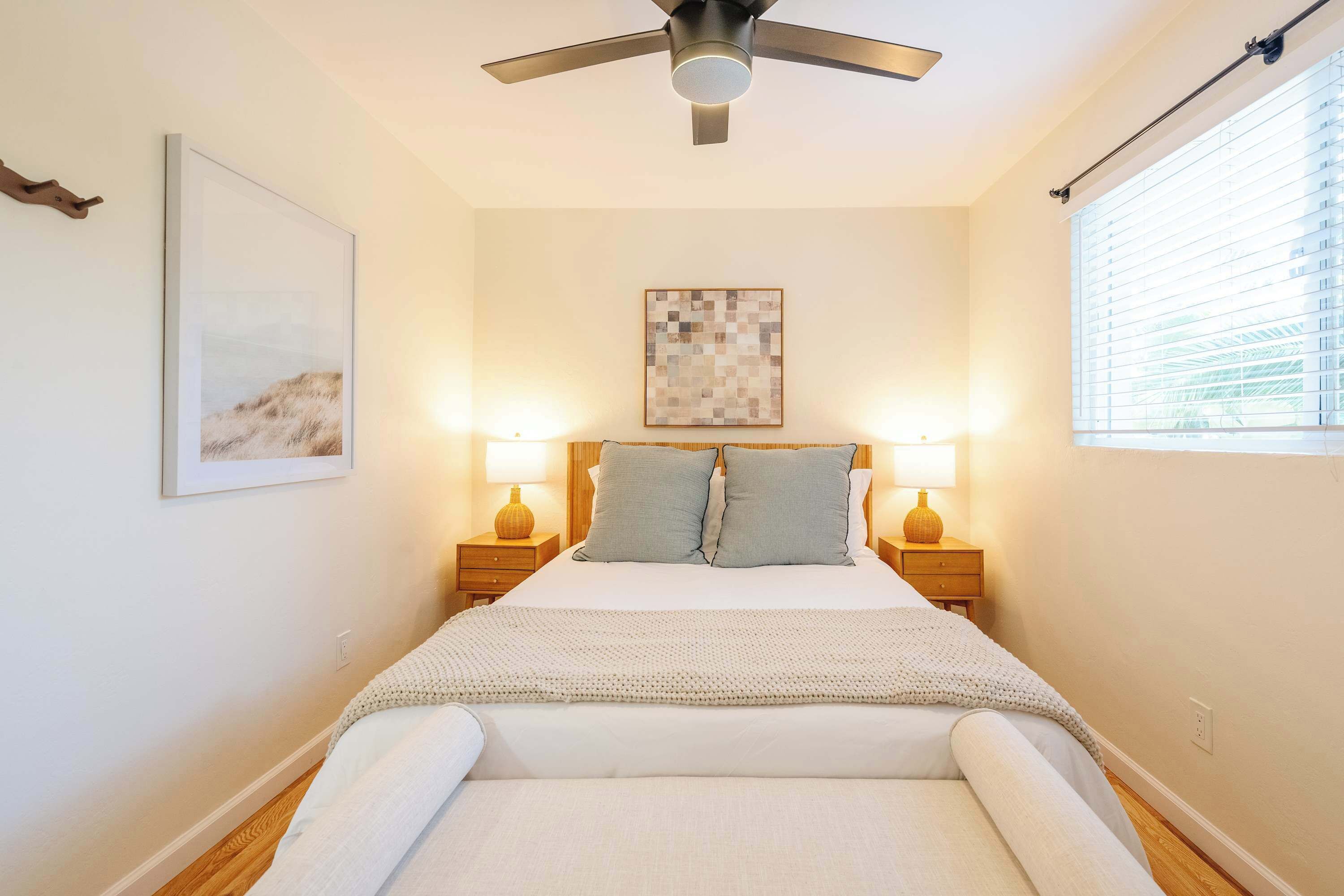 Une chambre à coucher avec un lit, deux lampes de chevet, un ventilateur de plafond, une fenêtre avec des stores et un tableau encadré au mur.