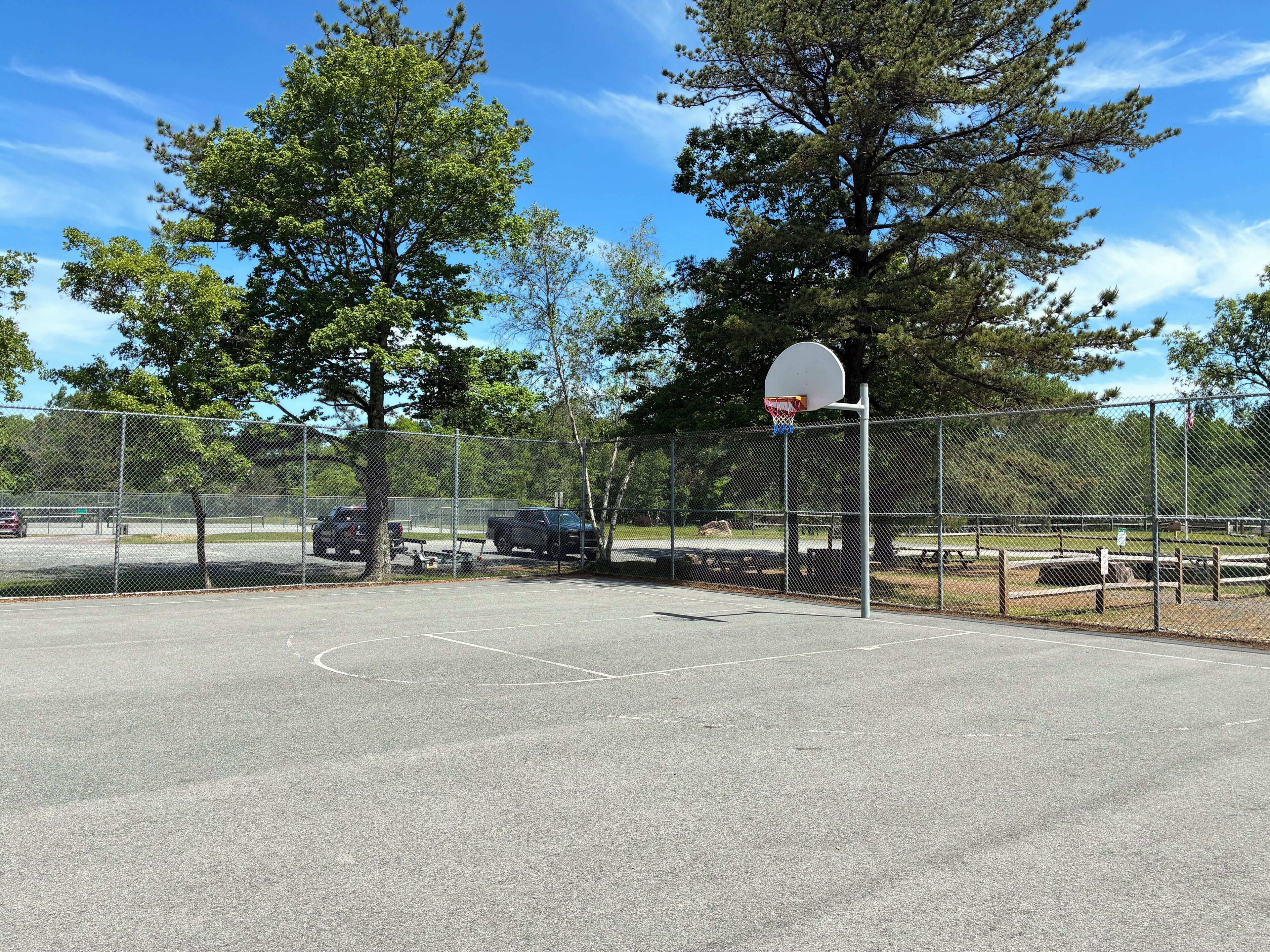 Un terrain de basket en plein air, entouré d’une clôture en treillis métallique, avec des arbres et un ciel dégagé.