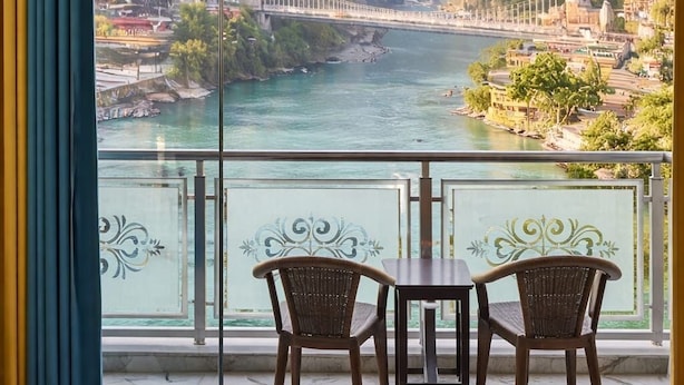 Uma varanda com vista para um rio, montanhas e uma ponte. Há duas cadeiras de vime e uma mesa pequena.