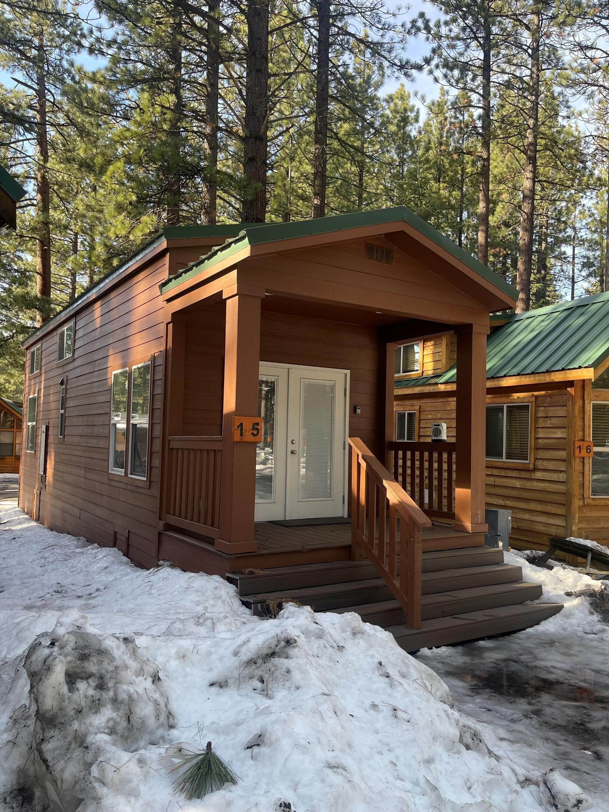 A small wooden cabin with a porch, numbered 15, surrounded by snow and pine trees.