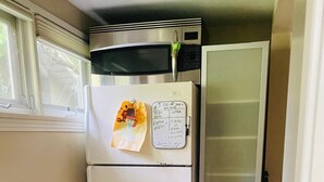 Eine kompakte Küche mit einem weißen Kühlschrank, einer Mikrowelle und einem Gefriertruhe. Links befindet sich ein Fenster und rechts eine Tür.