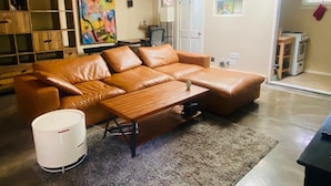 Ein Wohnzimmer mit braunem Ledersofa, einem Holztisch, einem Teppich, einer weißen Mülleimer und einer Küchenzeile mit Herd.