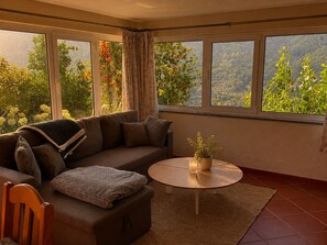 Een kamer met een bank, een kleine ronde tafel en uitzicht op groen door grote ramen.
