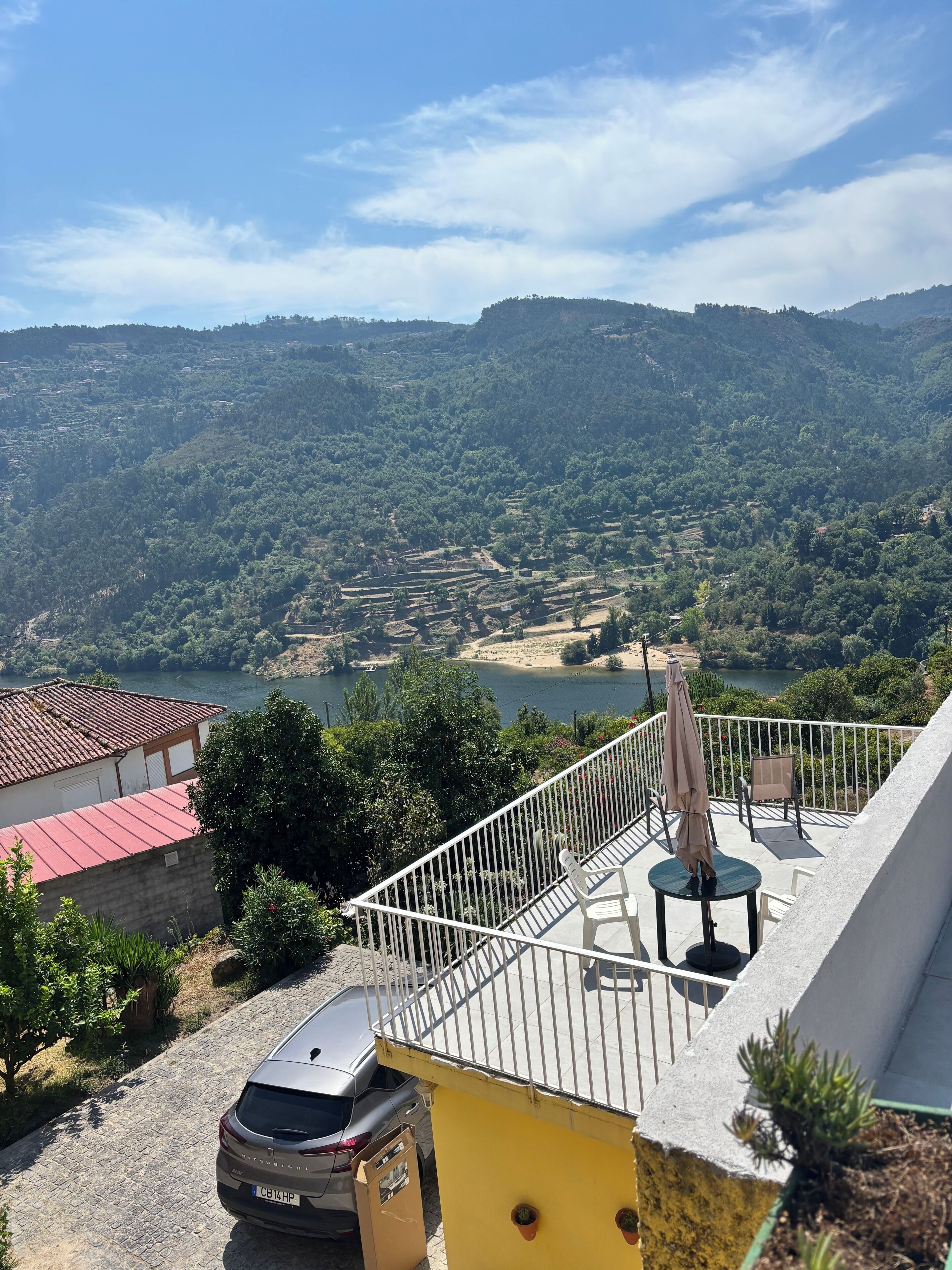 A balcony with a view of a lake and hills, a parked car, and a building with a red roof.