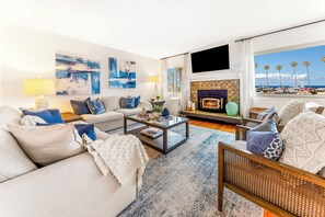 A living room with a fireplace, a sofa, a coffee table, and a view of the beach. - La Jolla Shores! Beach Across Street+Walk Everywhere (San Diego)