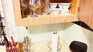 A kitchen with a wooden shelf holding mugs and glasses, a knife block, a paper towel holder, and a trash bin.