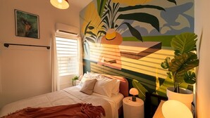 Ein Schlafzimmer mit einem Bett, Nachttisch, Lampe, Topfpflanze und einer Wand mit einem tropischen Wandbild.