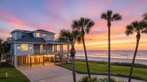 A two-story beachfront house with a balcony, palm trees, and a view of the ocean at sunset. - Beachfront 5BR | Hot Tub & Fire Pit | Game Room (Galveston)