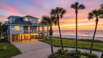 Une maison de deux étages située en bord de mer, dotée d’un balcon, de palmiers et offrant une vue sur l’océan au coucher du soleil.