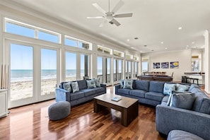 A spacious living room with large windows, a ceiling fan, and a view of the beach. - Beachfront 5BR | Hot Tub & Fire Pit | Game Room (Galveston)