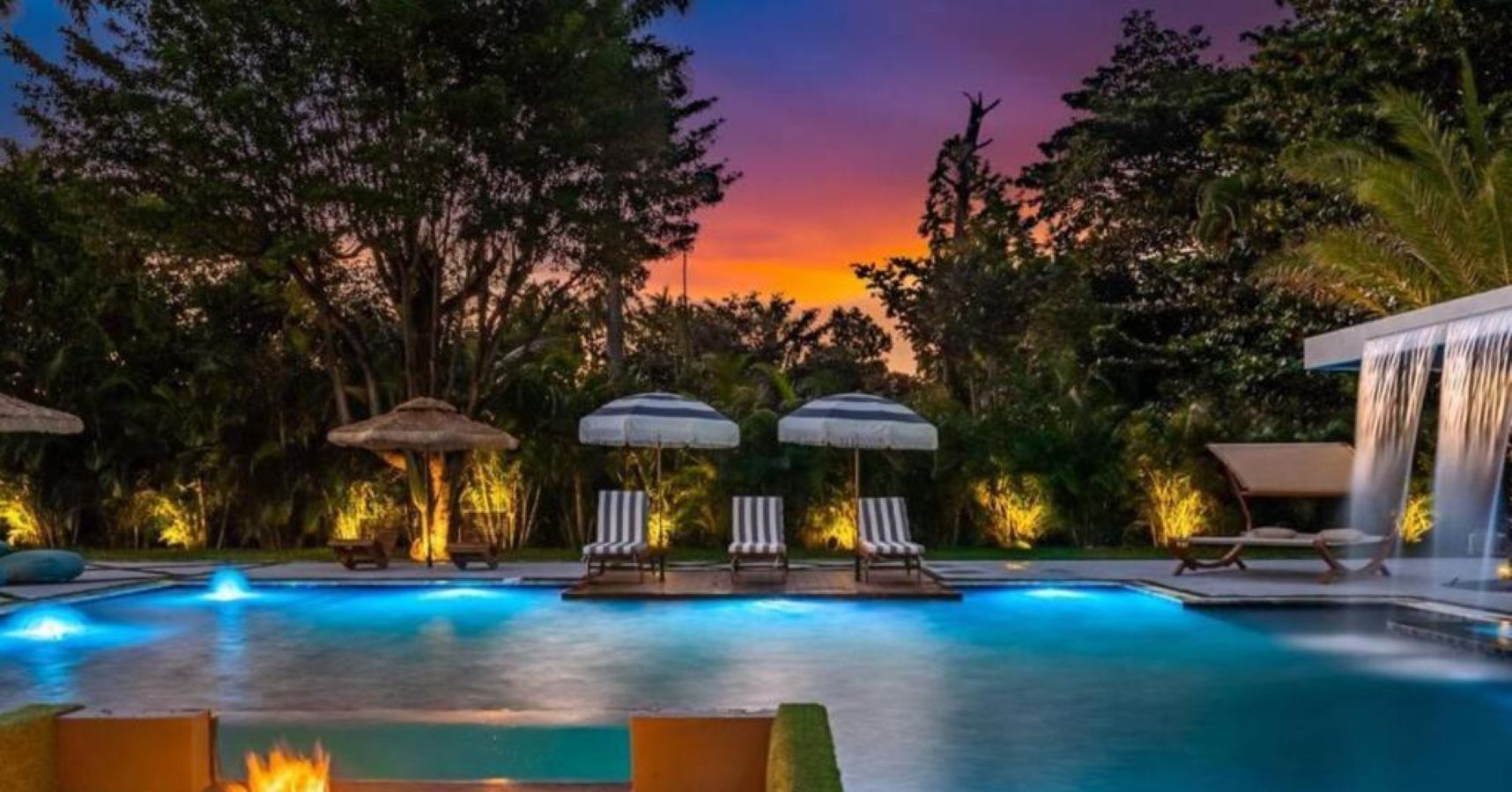 A swimming pool with lounge chairs and umbrellas, surrounded by trees and a sunset.