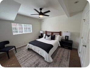 Ein Schlafzimmer mit Bett, Deckenventilator, Nachttisch, Stuhl und Fenster.