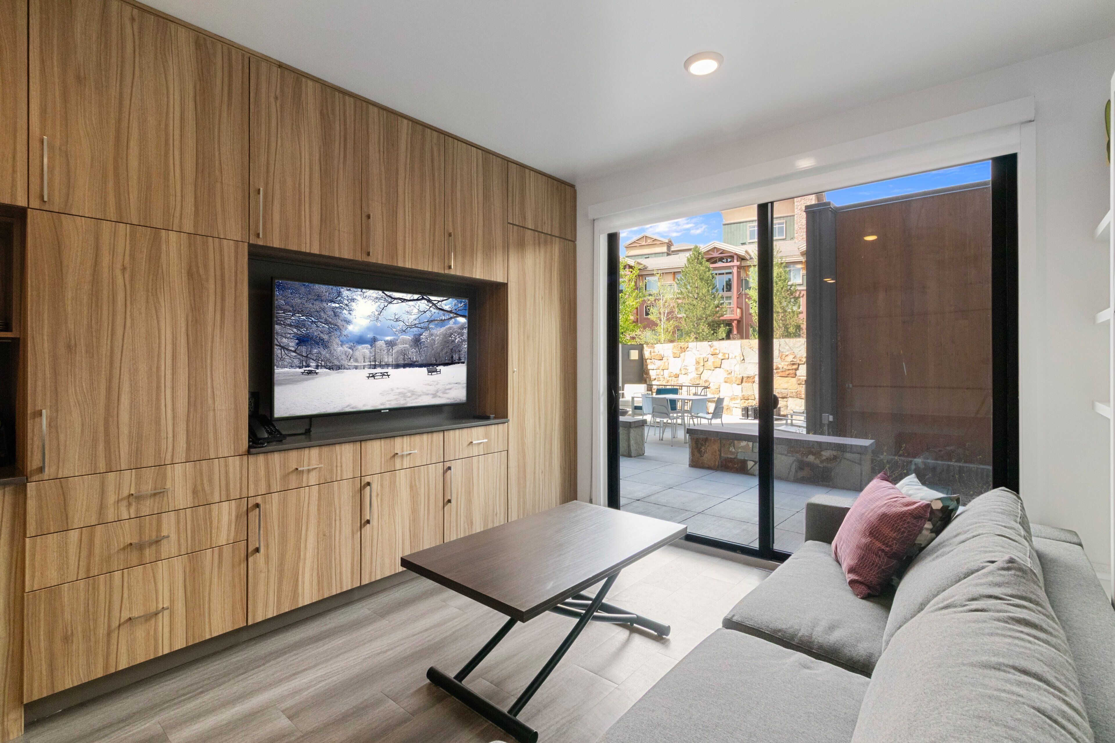 A modern living room with a TV mounted on a wooden wall, a sofa, a coffee table, and a sliding glass door leading to an outdoor patio.