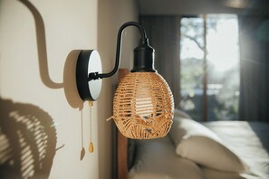Une lampe murale dotée d’un abat-jour tissé et d’une frange décorative, un lit avec une literie blanche et une fenêtre laissant entrer la lumière naturelle.