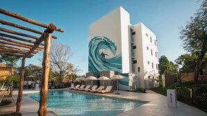 Ein Schwimmbad mit Liegestühlen, einer Pergola und einem Gebäude mit Wandgemälde im Wellenstil.