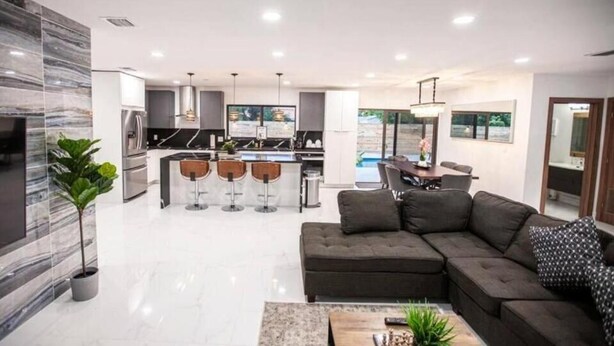 A modern kitchen with bar stools, a dining area with a table and chairs, a large sofa, and a TV. - Villa Dubai by Glam Homes (Ojus)