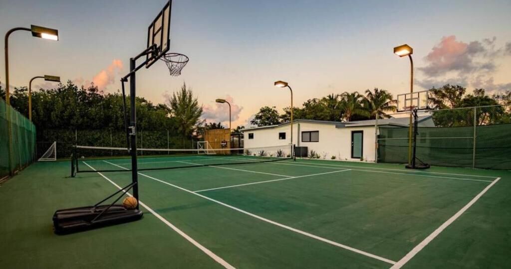 An outdoor tennis court with a basketball hoop, a small building, and street lamps.