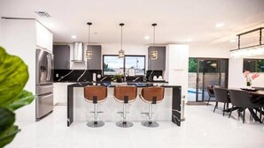 A modern kitchen with a central island, bar stools, and a dining area with chairs. - Villa Dubai by Glam Homes (Ojus)