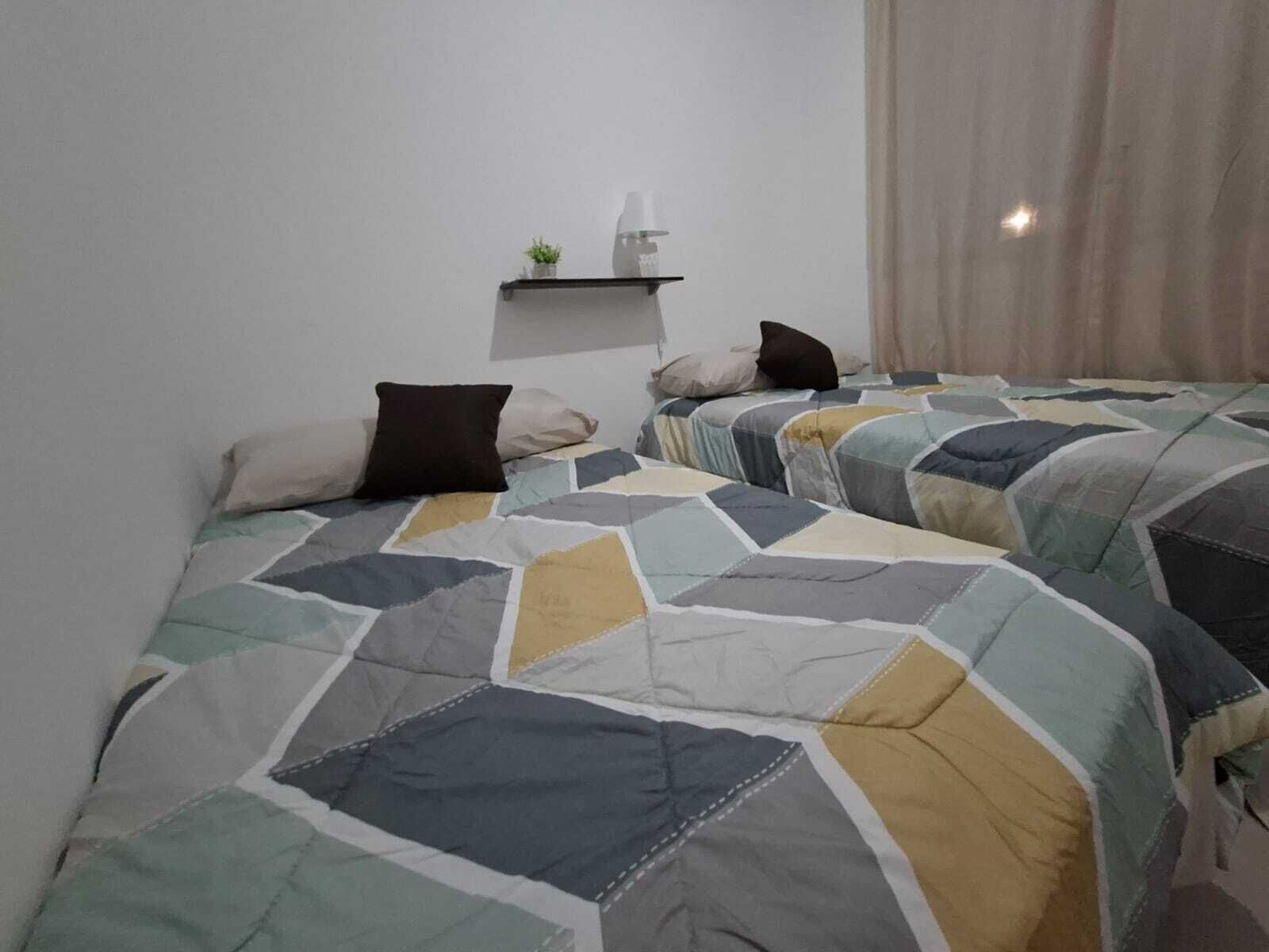 Een slaapkamer met twee bedden, elk voorzien van een bedovertrek met een geometrisch patroon. Er is een klein plankje met een plant en een flesje, en een raam met gordijnen.