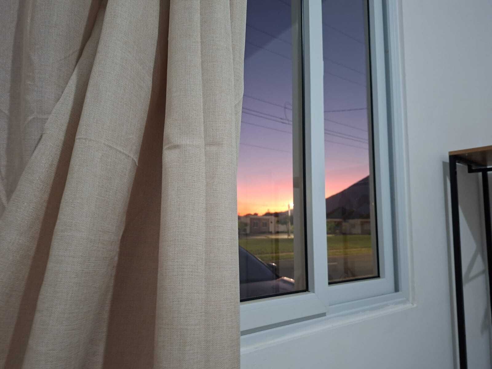 Finestra amb vista al capvespre i una muntanya, cortines mitjanament desplegades, i marc de metall negre.