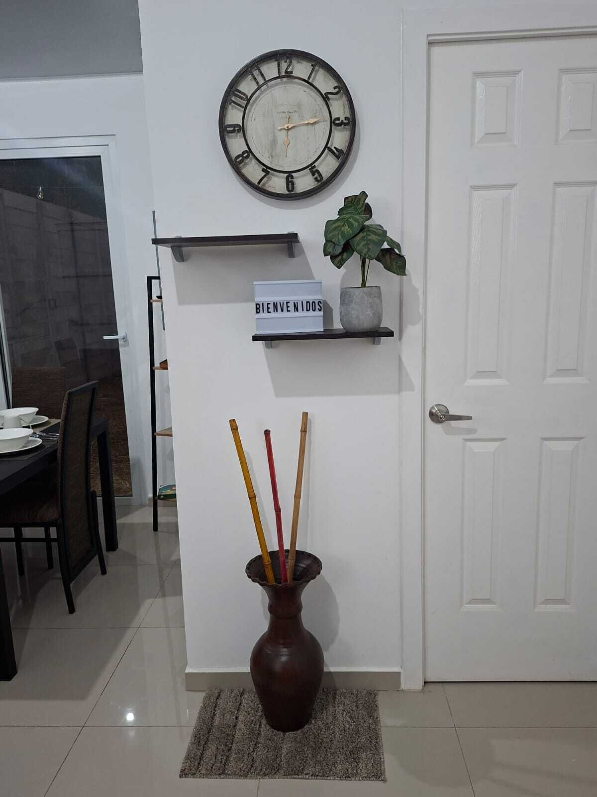 Ein Zimmer mit einer Wanduhr, einem Willkommensschild, einer Topfpflanze und einer Vase mit Ästen.