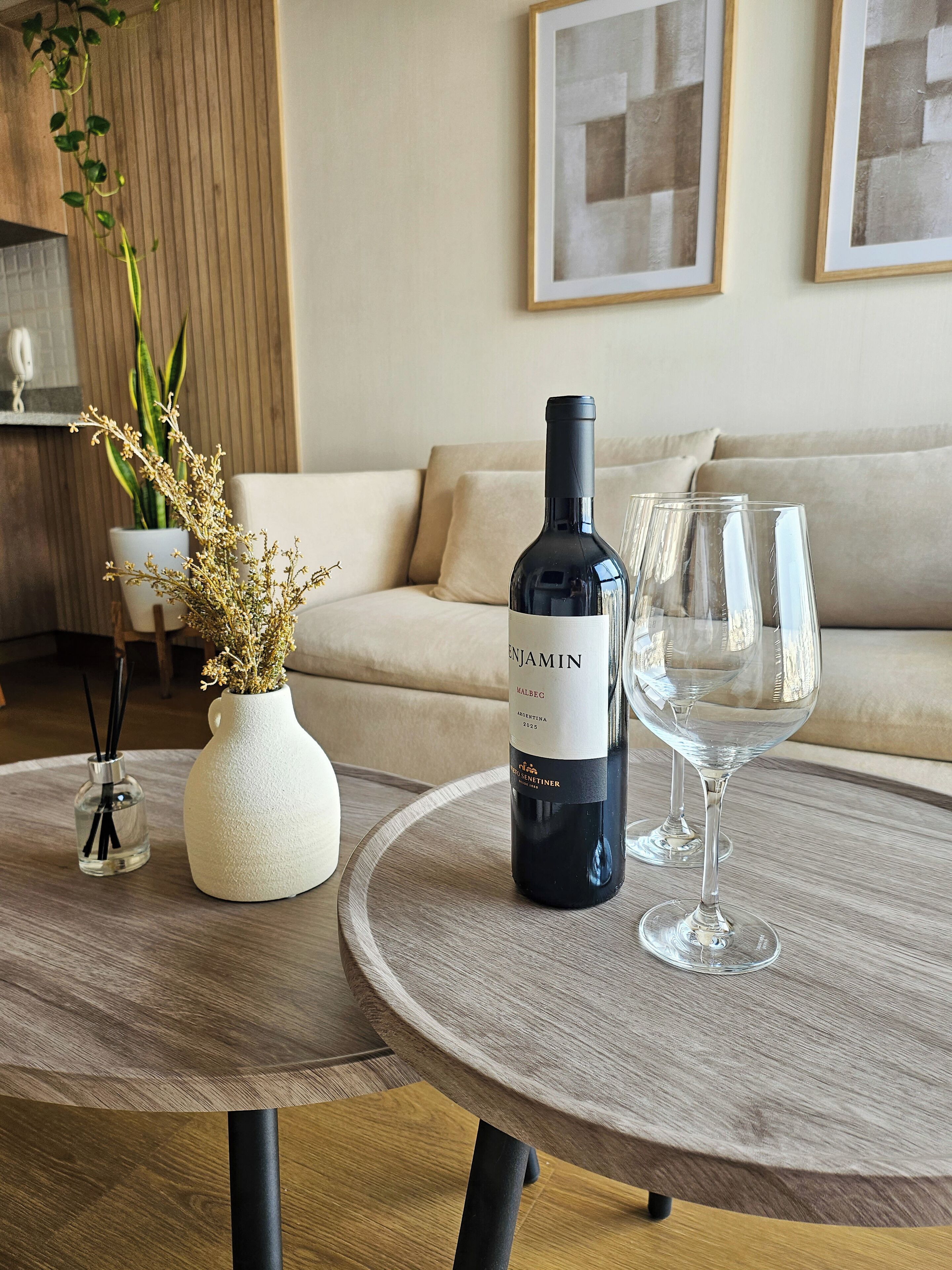 Une bouteille de vin et un verre sur une table ronde, un vase avec des fleurs séchées et un canapé en arrière-plan.