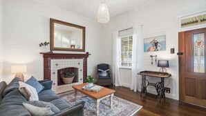 A living room with a fireplace, sofa, coffee table, and a painting on the wall. - Pip's Cottage - Pets Friendly, Cafes, Bars, Jetty Beach (Coffs Harbour)